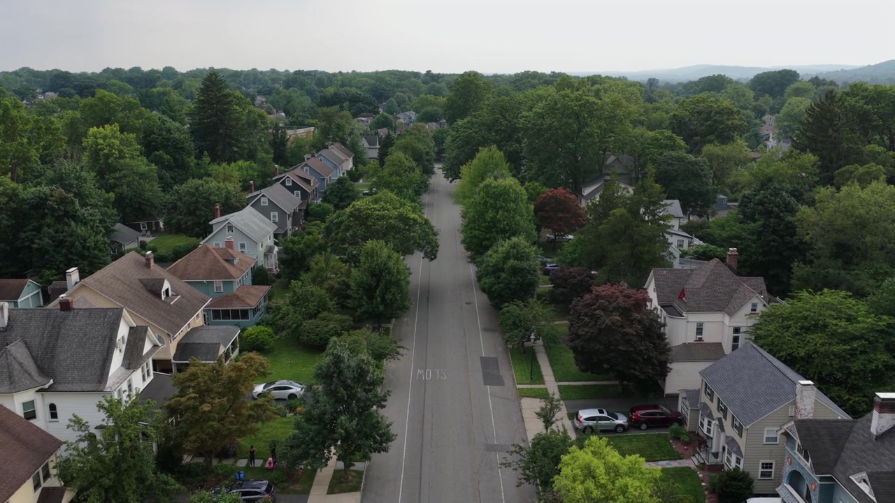 Strada suburbana alberata in un quartiere del New Jersey che mostra case in stile coloniale con tetti a capanna ripidi, portici avvolgenti, comignoli in mattoni e prati verdi ben curati. Ripresa aerea in discesa con drone.