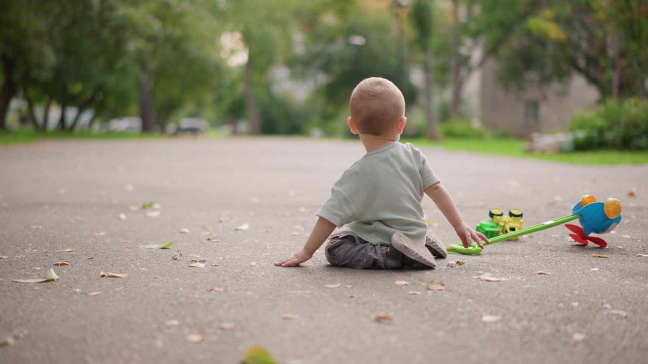 Bambino piccolo seduto sul marciapiede con un camioncino giocattolo, allunga la mano ed esplora le ruote colorate, una scena di gioco spontanea all'aperto che mostra curiosità, sviluppo motorio precoce e routine genitoriale.