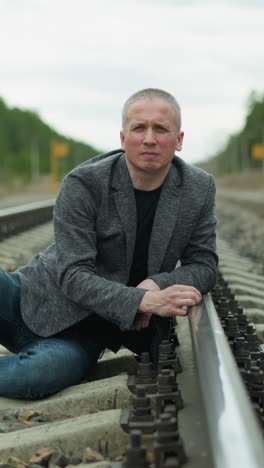 a man in a grey blazer and blue jeans sitting thoughtfully on a railway track in a rural area with a forest background, the man appears contemplative, resting on the tracks