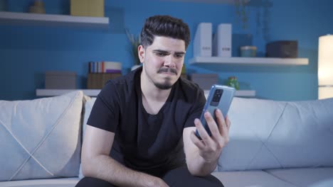 Young-man-looking-at-his-smartphone-at-home-feeling-emotional-and-upset.