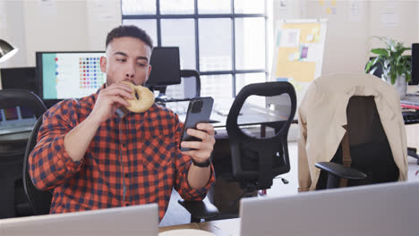 hombre biracial sentado en un escritorio, comiendo, mirando su teléfono inteligente y sonriendo, en cámara lenta