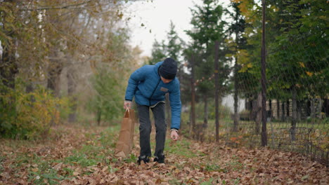 little boy in blue jacket and beanie bends down to pick rusty leaf from ground and places it in paper bag, with bar fence and building in background, surrounded by autumn leaves