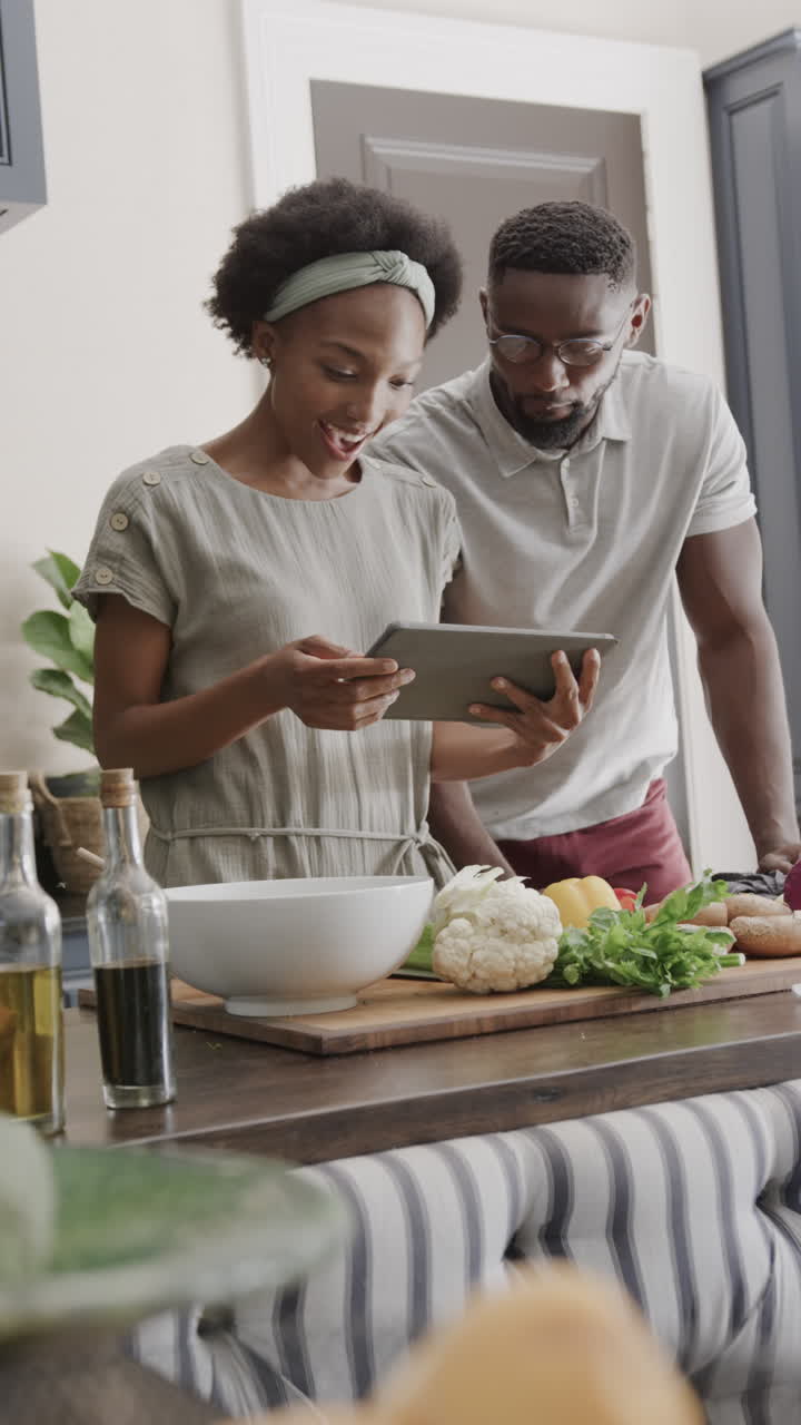 Vertical video of happy african american couple cooking and using tablet in kitchen in slow motion