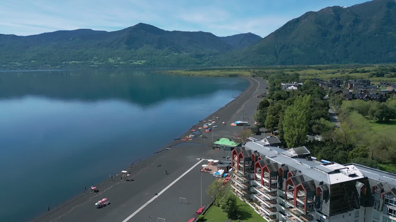 Panoramic drone shot of a peaceful lake with black sand beach, modern buildings, and mountains in the distance, filmed on a sunny day. Pucon Chile