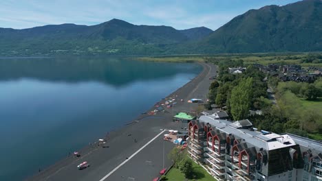 panoramic drone shot of a peaceful lake with black sand beach, modern buildings, and mountains in the distance, filmed on a sunny day. pucon chile