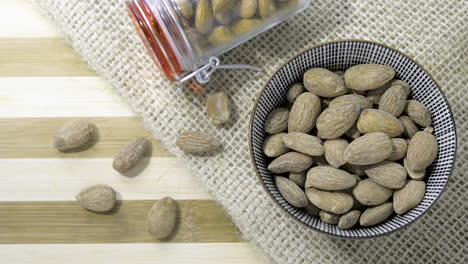 Top-down-panning-shot-across-striped-wooden-table,-Glass-jar-ceramic-bowl-filled-with-golden-almonds-set-on-striped-wooden-table-hessian-cloth