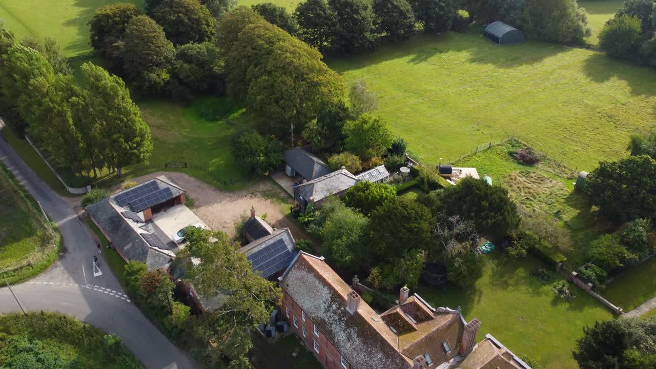 Drone moves forward above fields, roads, and a farmhouse with solar panels, surrounded by trees and a flock of sheep in East Sussex