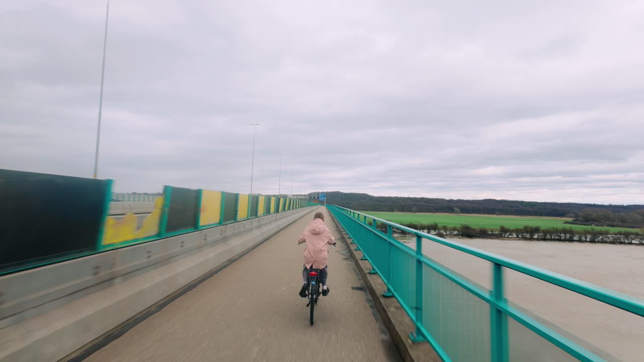 Vista posteriore di una ragazza giovane in bicicletta su una pista pavimentata di un ponte con vista su campi verdi e un fiume nella zona di Renkum