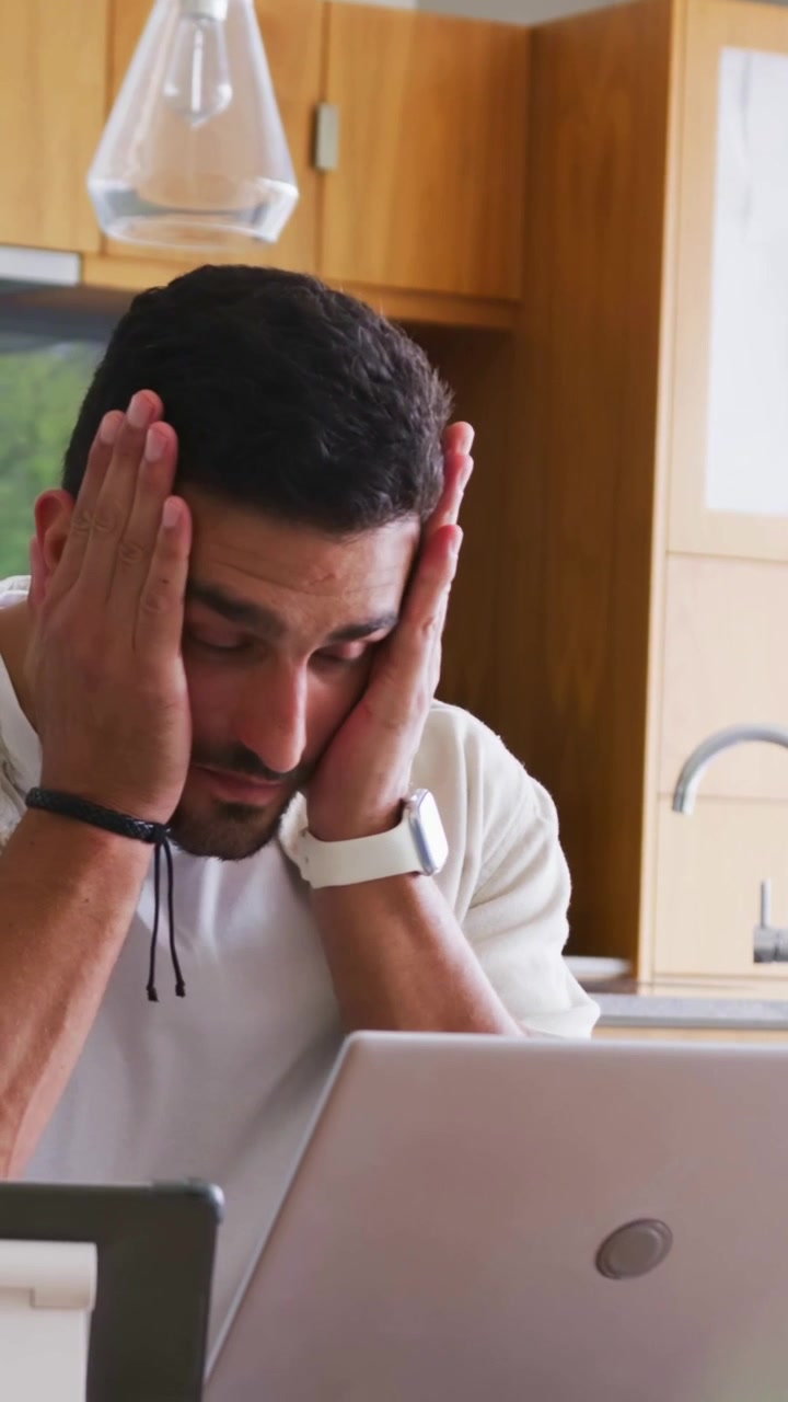 Video vertical: Hombre con jersey mira su portátil, apretándose las mejillas y frotándose la mandíbula en la encimera de la cocina.