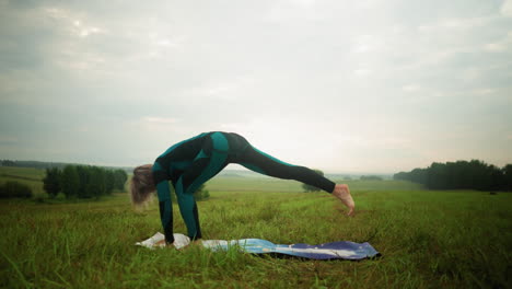 woman outdoor in vast grassy field on yoga mat practicing low lunge pose, bending forward with her hands touching the mat, under a cloudy sky, surrounded by trees on the horizon