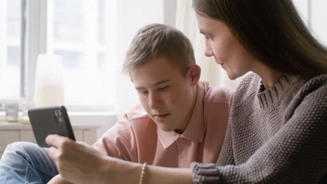 Nahaufnahme-Eines-Jungen-Mit-Down-Syndrom-Und-Seiner-Mutter,-Die-Etwas-Auf-Dem-Smartphone-Beobachtet,-Das-Zu-Hause-Auf-Dem-Bett-Im-Schlafzimmer-Liegt