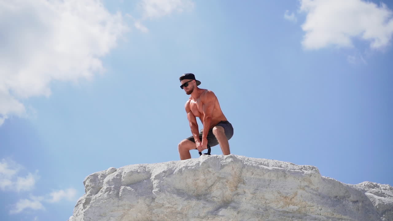 Healthy athlete swinging kettlebell outdoors. Fitness training man lifting heavy kettlebell weight during workout on the top of white mountain.