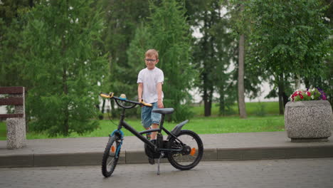 a young boy walks playfully toward a parked bicycle and moves its handlebar, he is wearing glasses, a white shirt, denim shorts, and sandals, with a green park setting in the background