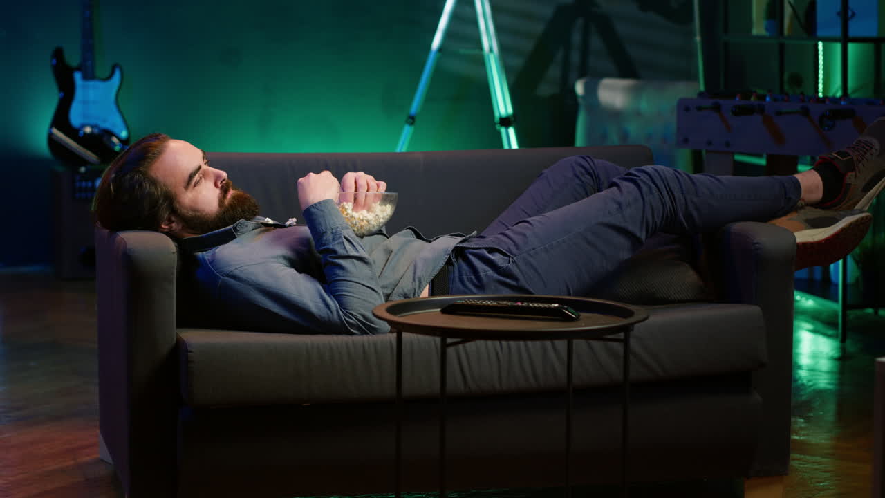 Bored man procrastinating at home, laying down on couch, watching TV