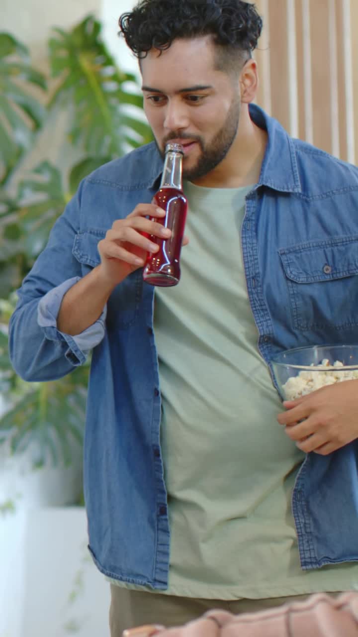 Video verticale: Uomo sorridente con camicia di jeans che alza una bottiglia, sorseggia e tiene in mano popcorn in soggiorno.