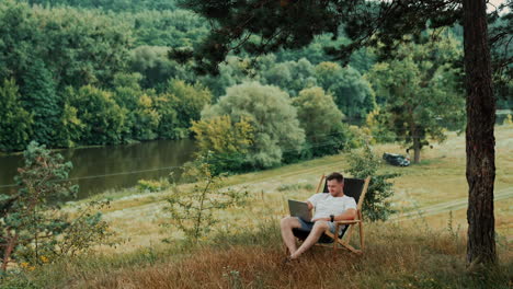remote work in the nature away from the office. caucasian smiling man is in folding chair holding his laptop.