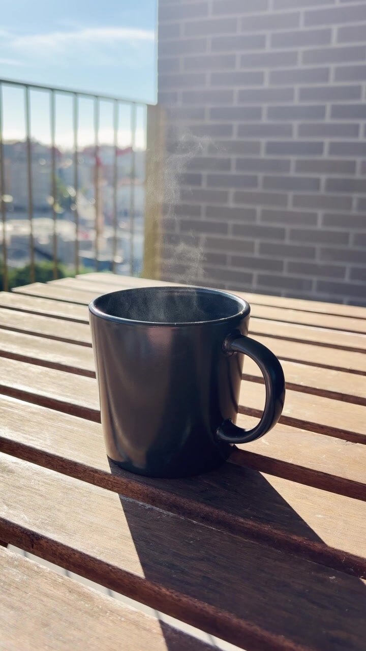 La luce del sole del mattino proietta lunghe ombre da una tazza nera di caffè caldo e fumante appoggiata su un tavolo di legno su un balcone in città, creando un inizio di giornata sereno e rilassante