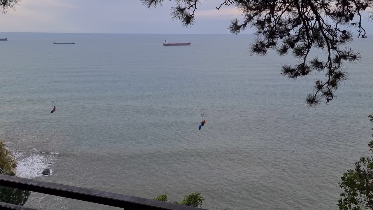 two person are taking zip line ride at Batumi botanical garden and on background we can see oil tankers in black sea.