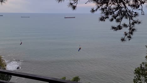 two person are taking zip line ride at batumi botanical garden and on background we can see oil tankers in black sea.