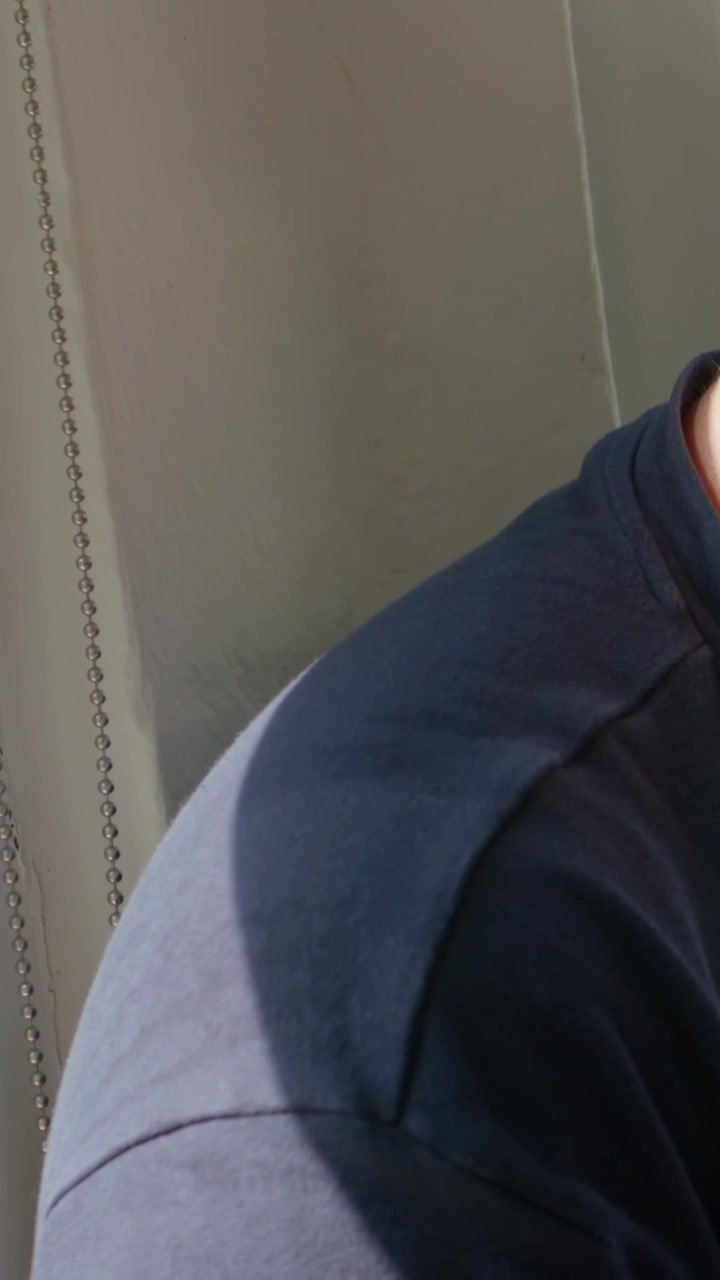 Video vertical: Vista panorámica mostrando a un hombre con camiseta azul marino revelando el brazo junto a la ventana, con una cadena y espacio para texto.