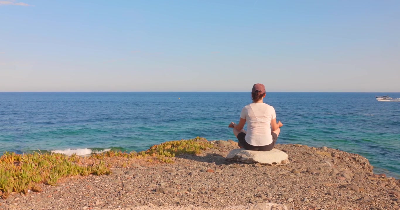 Persona che medita in posizione yoga seduta su una riva rocciosa di fronte a un mare blu calmo.