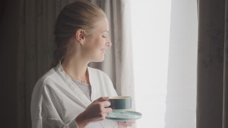 woman wearing bathrobe on hotel or spa break standing by open curtains with hot drink