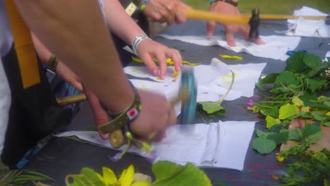 group of adults and children are bashing hitting leaves with hammer placed under a piece of cloth to create artistic piece out of it close up on hands flowers on table family are on music festival