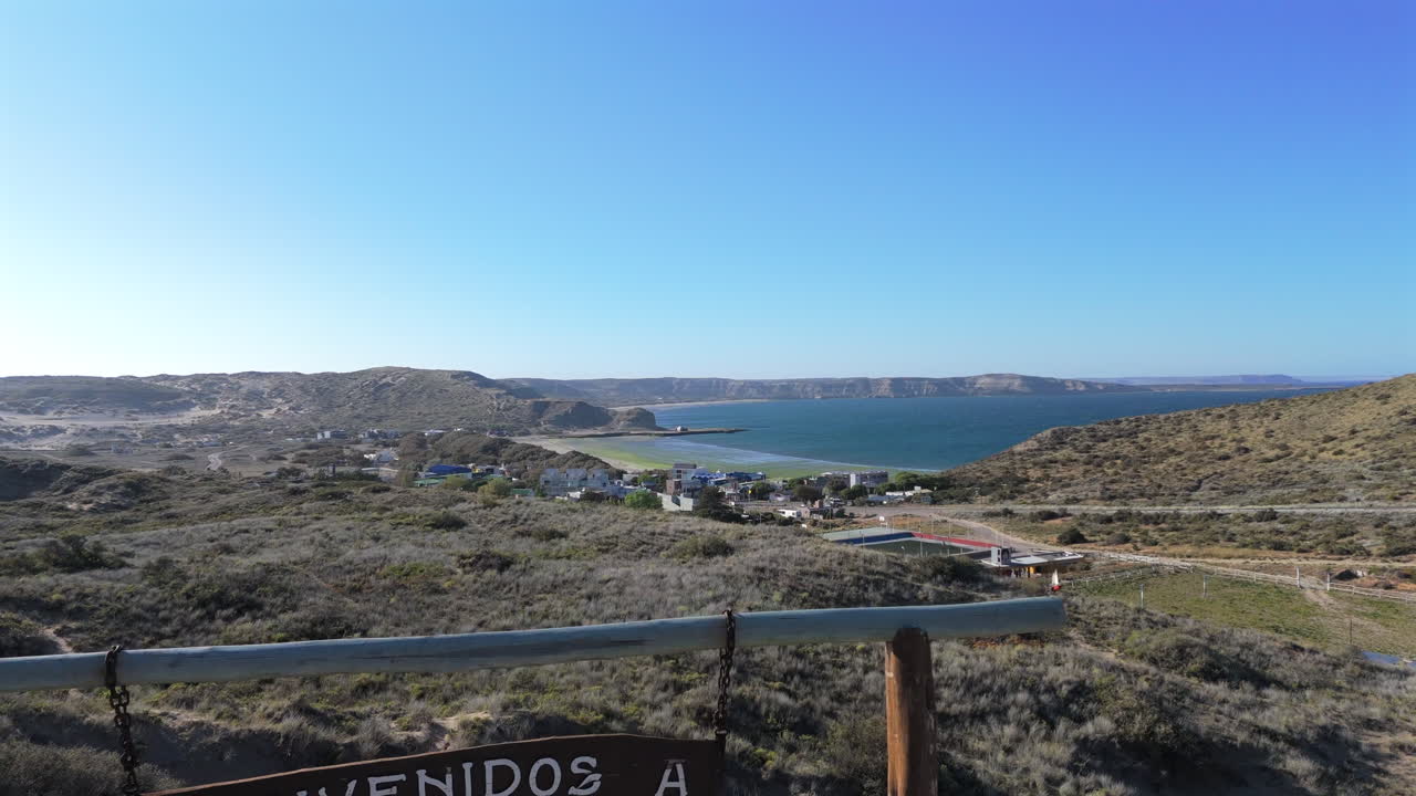 Low altitude drone shot of Puerto Pirámides welcome sign, with road, cityscape, and ocean backdrop. Beautiful blue afternoon sky. Shot on 4K at 60fps.
