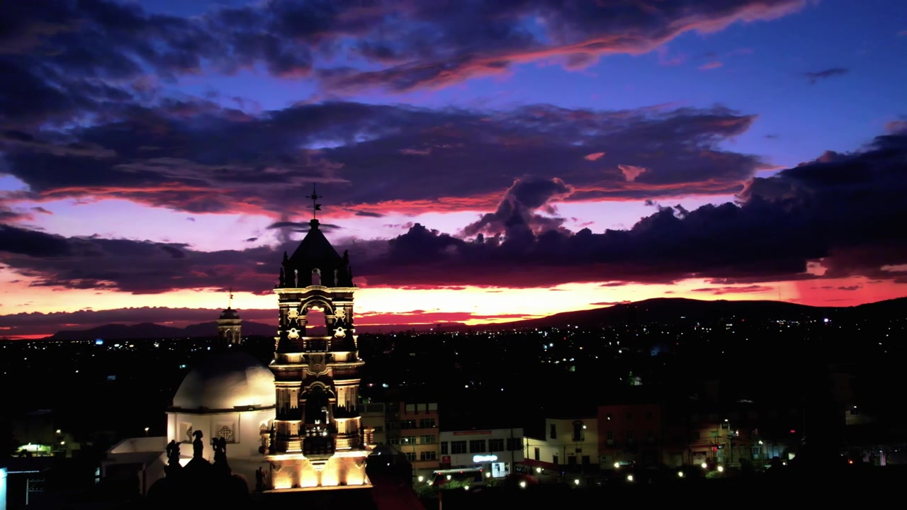 Vista panoramica di Irapuato, Guanajuato al tramonto, con una torre campanaria illuminata, luci urbane, colline circostanti e nuvole drammatiche sulla città nel Messico centrale.