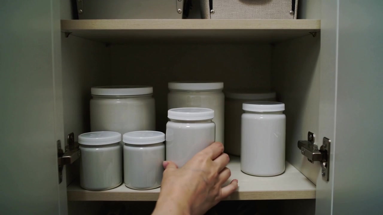 Un braccio nudo si allunga verso una maniglia nera, aprendo un mobile da cucina, rimuovendo un contenitore alto e sistemando.
