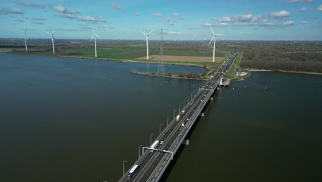 El-Dron-Vuela-Alto-Sobre-Un-Río-Y-Un-Puente-Para-El-Tráfico,-Turbinas-Eólicas-En-El-Horizonte