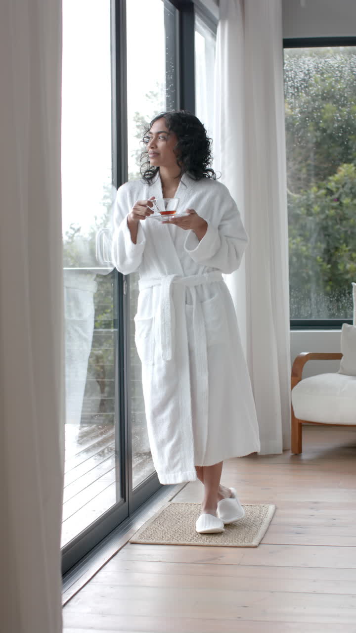 Vertical video of biracial woman wearing bathrobe drinking tea at home, slow motion