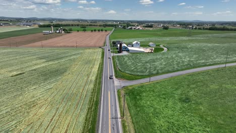 traffic scene on american rural suburb intersection between farmstead and agricultural farm fields. sunny day in spring season. aerial approaching wide shot.