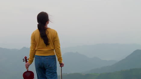 Mujer-Con-Violín-Caminando-Sobre-Una-Colina-En-Un-Clima-Nublado-Con-Fondo-De-Montaña