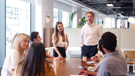 hombre de negocios y mujer de negocios dirigiéndose a un grupo de jóvenes candidatos sentados alrededor de una mesa y colaborando en una tarea en el día de evaluación de reclutamiento de graduados
