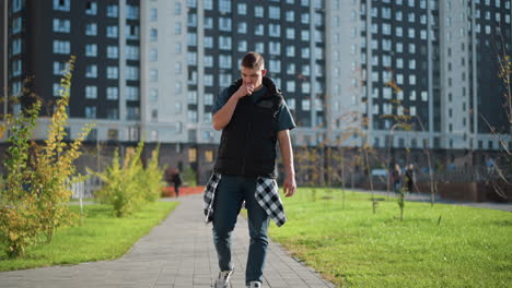 young man in puffer vest and checkered shirt strolls along paved path inhaling from vape device then coughs into hand on chest as thick smoke puffs out near tall apartment buildings