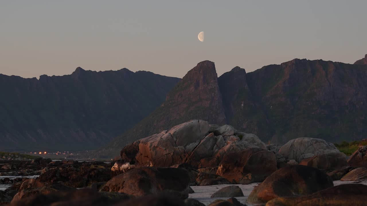 A half moon rises behind the mountains, illuminating a beach scattered with smooth, round stones. A serene and cinematic night scene perfect for nature footage