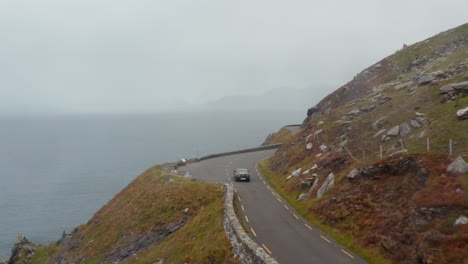 Vorwärtsverfolgung-Eines-Schwarzen-Autos,-Das-Auf-Einer-Schmalen,-Exponierten-Küstenstraße-Hoch-Auf-Einer-Klippe-Fährt.-Dunstiger-Blick-Auf-Meer-Und-Hügel.-Irland