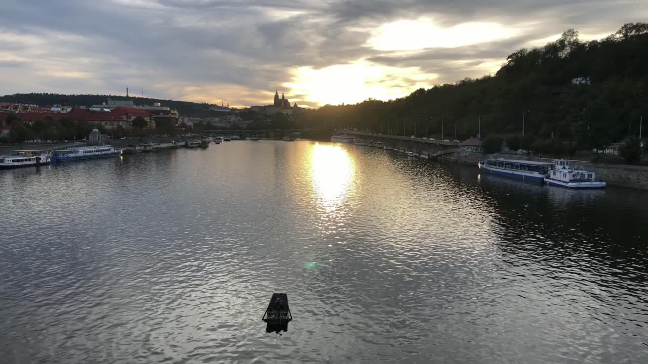 Qualcuno siede all'interno di una barca a remi che galleggia da sola nel mezzo del fiume Moldava mentre il sole della sera tramonta dietro il Castello di Praga sullo sfondo