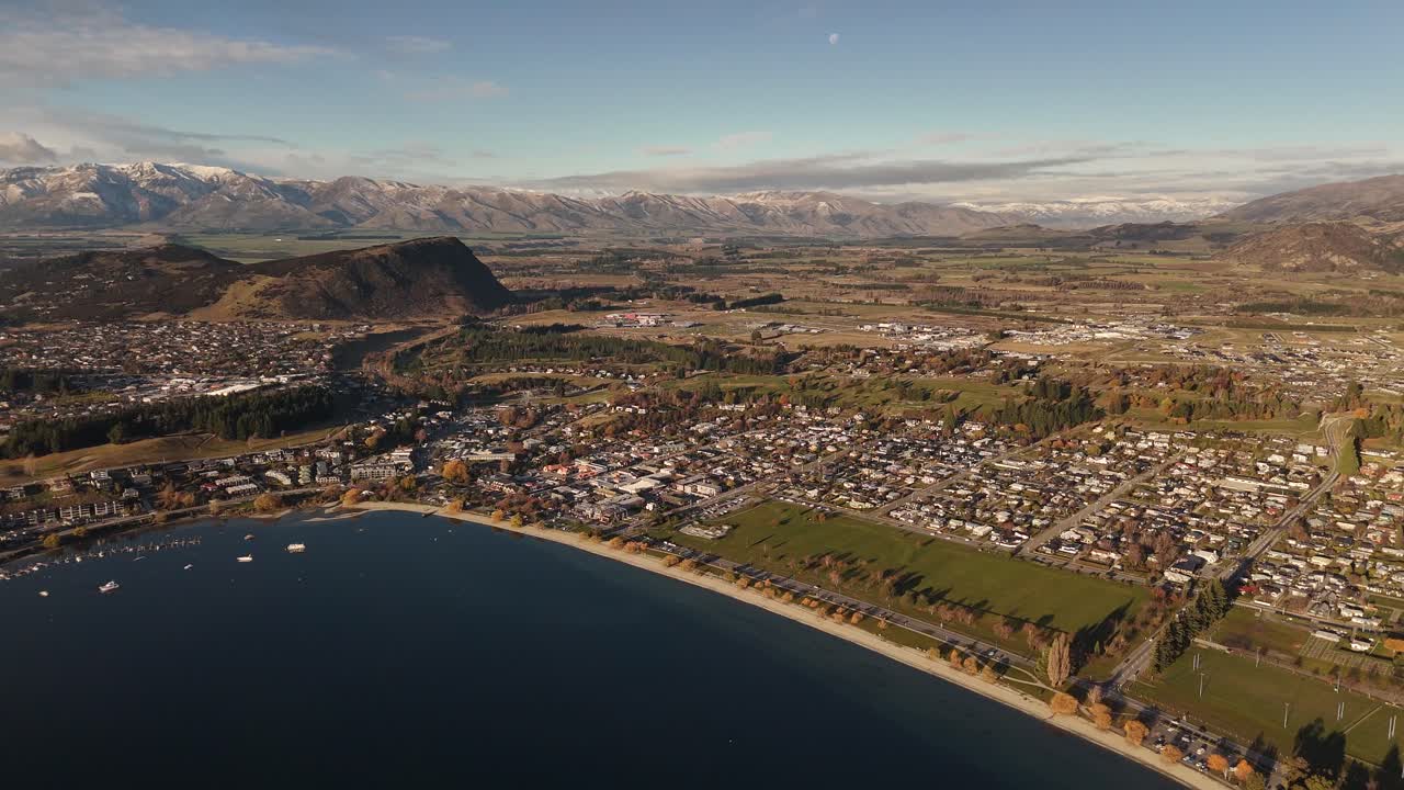 Breathtaking 4K 60fps panoramic shot of Wanaka town and Lake Wanaka during a spectacular sunset. The golden hour light creates a warm, beautiful glow over the entire landscape