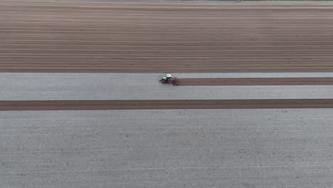 drone tracks tractor leveling soil in slow motion, carving uniform dark seed rows beside light-bleached furrows across an expansive field under warm dusk light, horizon fading into soft evening tones