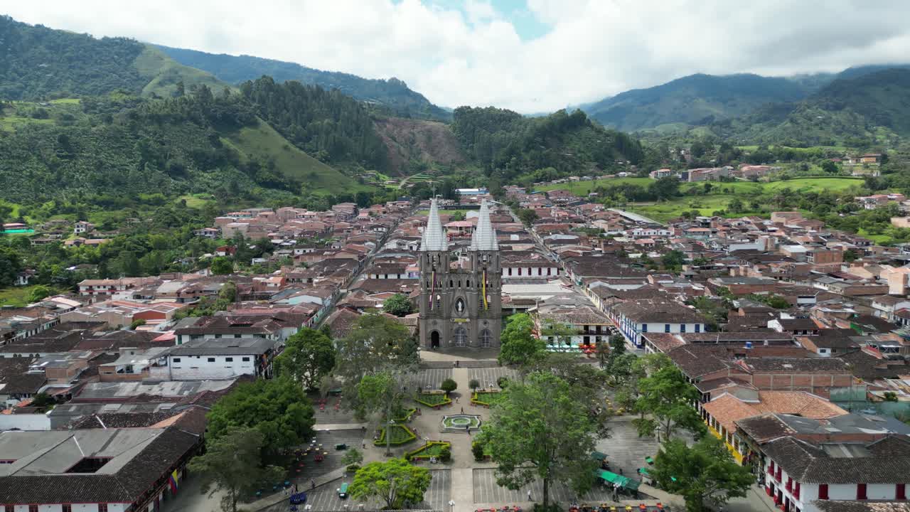 Aerial footage of the charming Andean town of Jardín in Colombia, featuring the vibrant central square, the church Basílica Menor de la Inmaculada Concepción and the green mountain landscape