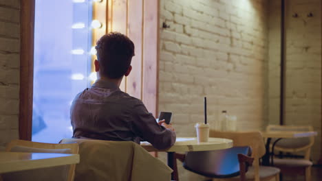 man seated alone in cozy cafe, facing frosty window while holding smartphone and coffee cup, wrapped in introspection. soft indoor lighting contrasts wintery outdoor tones, evoking mood of solitude