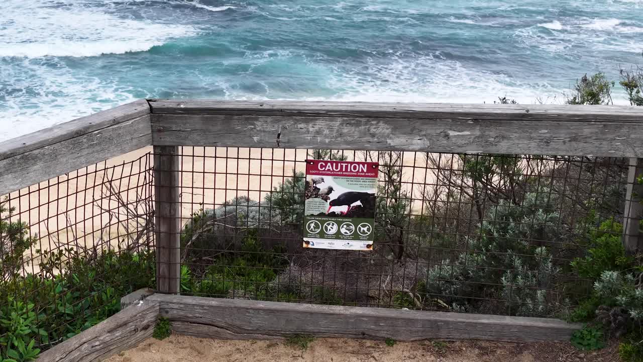 Camera tilts down from warning sign to sandy beach and turquoise waves under cloudy daylight
