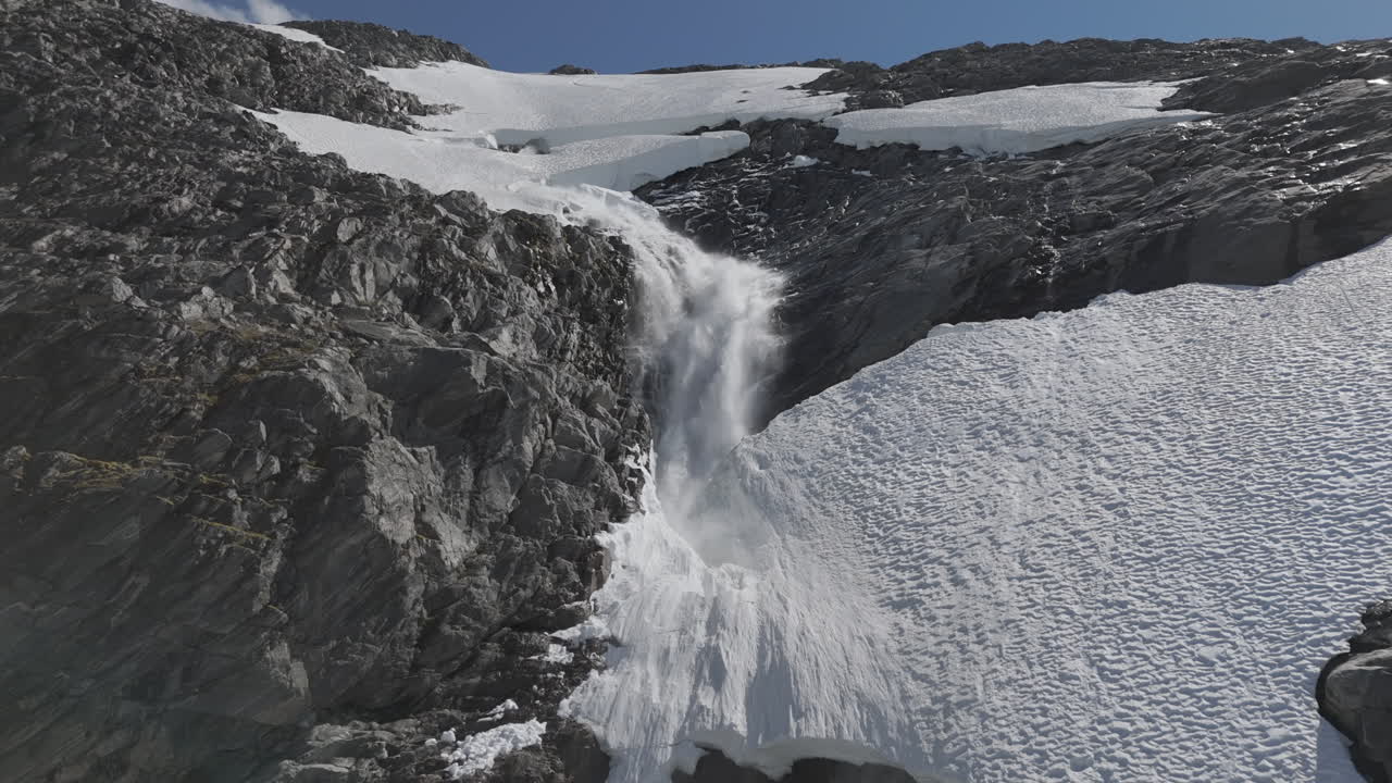 un avión no tripulado en cámara lenta volando alrededor de una poderosa cascada cerca del lago langvatnet en noruega cerca de strynefjellsveg rompiendo a través del hielo y la nieve en un día soleado rodeado de rocas brillantes