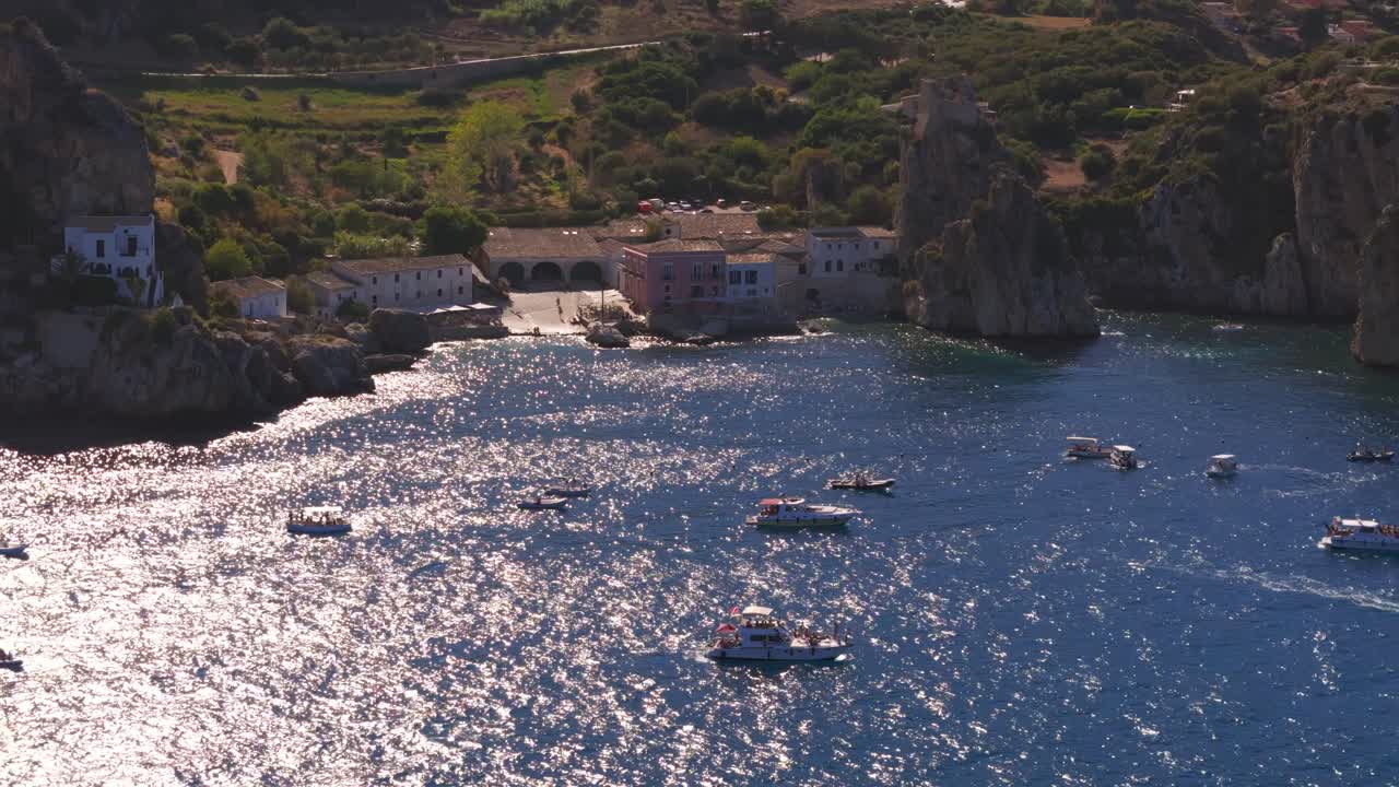 Aerial drone shot panning right above the calm blue waters at Tonnara di Scopello, Sicily, Italy, passing over numerous boats as the ocean shimmers under the golden sunlight during Italian summer