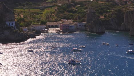 aerial drone shot panning right above the calm blue waters at tonnara di scopello, sicily, italy, passing over numerous boats as the ocean shimmers under the golden sunlight during italian summer