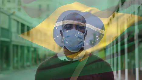 animation of flag of brazil waving over man wearing face mask during covid 19 pandemic