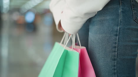 close-up of woman in white sweatshirt and jeans standing indoors holding mint and pink shopping bags, blurred background with colorful lights and mall setting