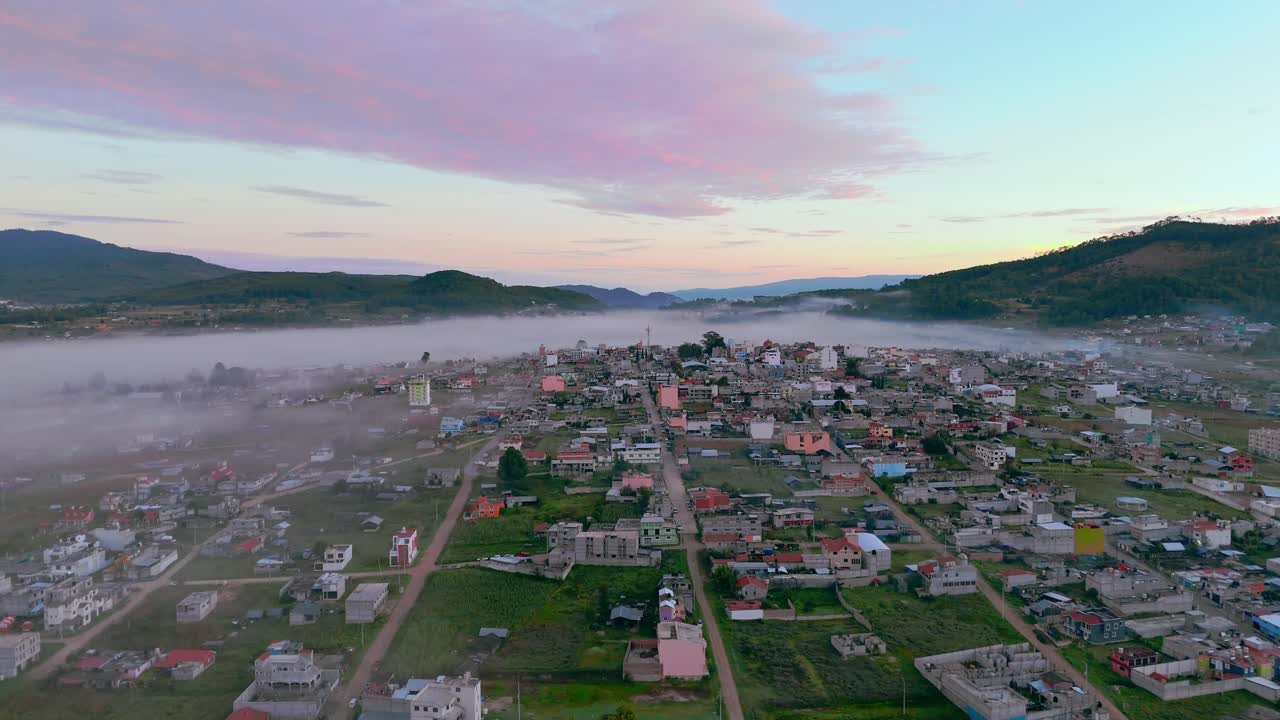 Aerial zoom in over Chalcatongo, Oaxaca. Early morning fog gently covers parts of the town with colorful buildings and mountains in the background.